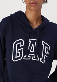 Kvinna i en marinblå GAP-hoodie med vit och marinblå text och snoddar, guldörhängen synliga.