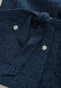 Nikot zils tvīda audums ar teksturētu virsmu, tajā ir sasiets jostas aizdare, divas priekšpuses kabatas un sudrabainas pogas mezgla formā.