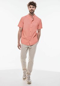 Lehká korálová košile s krátkým rukávem a mandarínským límcem, s button-up přední stranou, a béžové zúžené kalhoty. Model má na sobě béžové tenisky.