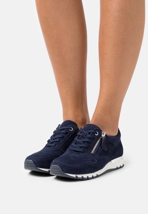 Legs wearing navy blue suede sneakers with laces and side zippers, standing on a white surface.