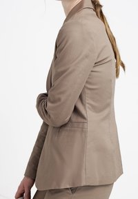 Hnědý šitý blazer vyrobený z hladké tkaniny, s jedním zadním rozParkem a mírně přiléhavým střihem. Žádné viditelné kovové prvky nebo ozdoby.