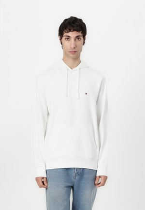 Weißer Pullover-Hoodie mit einer Kängurutasche, Kapuze mit Zugband und kleinem gesticktem Logo auf der linken Brust. Kombiniert mit blauen Jeans.