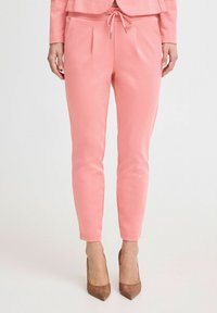 Rosa elastische Jogginghose mit Falten an der Vorderseite, kombiniert mit braunen spitzen High Heels. Glatte Textur und figurbetonte Form.