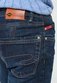Jeans en denim bleu foncé avec des coutures orange, une étiquette en cuir et deux poches arrière avec des accents en métal argenté.