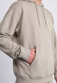 Beige kapucnis pulóver, elülső zsebből, húzózsinóros kapucnival és sárga kör alakú logóval. Puha anyag, bordázott mandzsetta és alj textúrával.