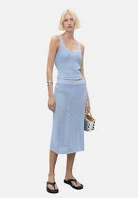 Top din tricot de culoare albastru deschis, fără mâneci, și fustă asortată, ambele cu un design texturat și cusătură deschisă. Modelul ține o geantă mică cu un model și poartă sandale negre.