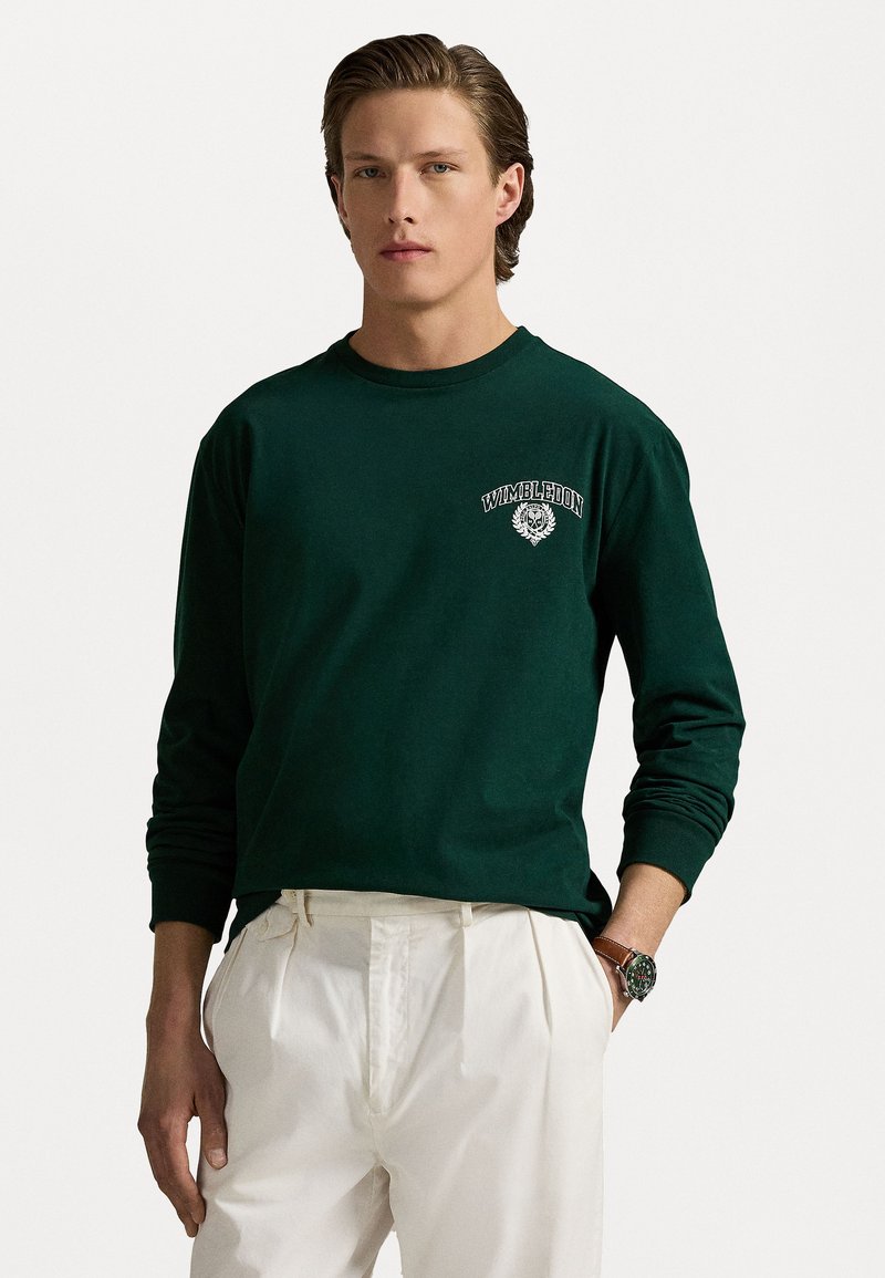 Groene T-shirt met lange mouwen van katoen, met een wit "Wimbledon" logo met een bloemenembleem op de linkerborst. Gecombineerd met witte broek.