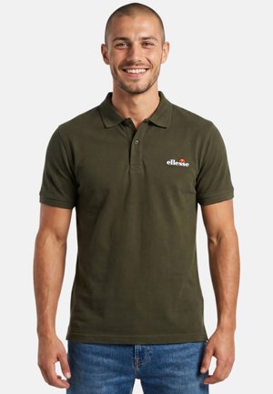 Homme souriant portant un polo ellesse vert olive et un jean bleu, debout devant un fond clair uni.