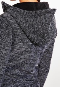 Personne portant un sweat à capuche en tricot gris foncé avec un tissu texturé et deux petites oreilles de chat triangulaires sur la capuche.