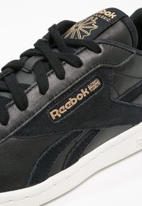 Baskets en cuir et daim noir avec des lacets noirs. Logo doré "Reebok Classic" et une semelle en caoutchouc blanche. Langue côtelée avec des emblèmes.