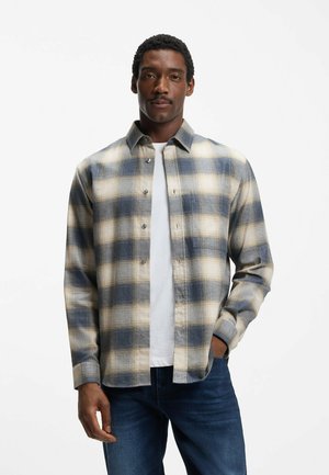 Mężczyzna w niebiesko-beżowo-kremowej koszuli w kratę na guziki, noszący białą koszulkę i ciemnoniebieskie dżinsy, stojący na tle białego tła.