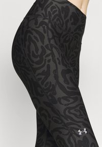 Primo piano di una persona che indossa leggings neri e grigi scuri con un motivo mimetico, dotati di un logo riflettente di Under Armour sulla coscia inferiore.