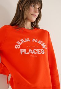 Femme portant un sweat-shirt orange vif avec le texte blanc « SEEK NEW PLACES COLLECTING STORIES », regardant vers le haut, main dans la poche.
