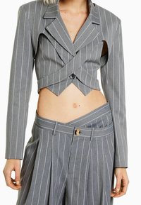 Femme portant un blazer cropped à rayures grises avec découpes et un pantalon assorti taille haute à rayures avec une ceinture asymétrique.