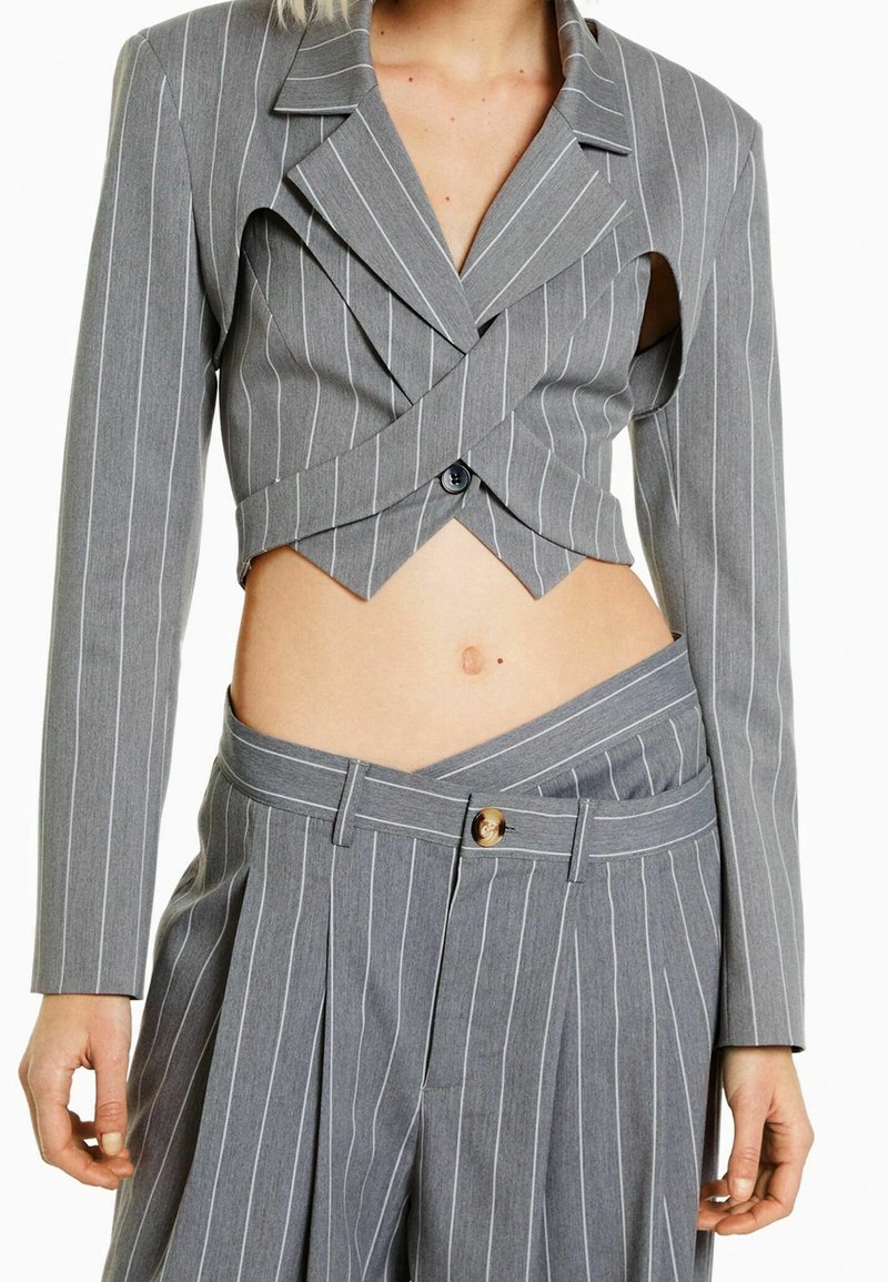 Femme portant un blazer cropped à rayures grises avec découpes et un pantalon assorti taille haute à rayures avec une ceinture asymétrique.