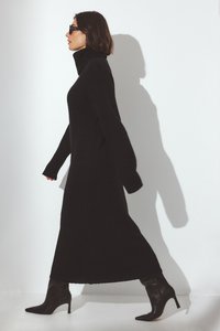 Pitkä musta neulemekko, jossa on korkea kaulus ja pitkät hihat, yhdistettynä teräväkärkisiin mustiin korkokenkiin. Silkkinen rakenne, minimalistinen muotoilu.