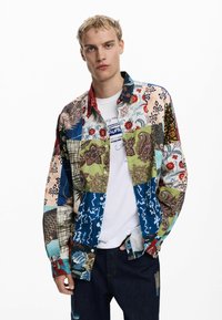 Camisa de patchwork multicolor que presenta varios patrones, incluidos diseños florales y geométricos. Lleva puesta sobre una camiseta blanca, con jeans de mezclilla.