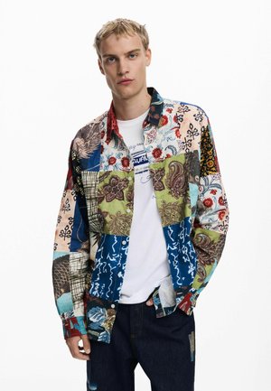 Multicolored patchwork shirt featuring various patterns, including floral and geometric designs. Worn over a white T-shirt, with denim jeans.