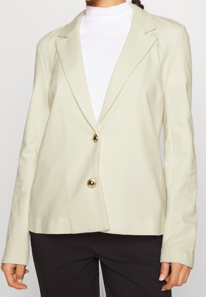 Femme portant un blazer crème avec deux boutons dorés, associé à un col roulé blanc et un pantalon noir, vue des épaules aux hanches.