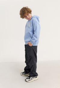 Gaiši zila kapuce ar priekšējo kabatu un izšūtu tekstu, saskaņota ar melniem baggy biksēm un masīvām melnbaltām kedām.