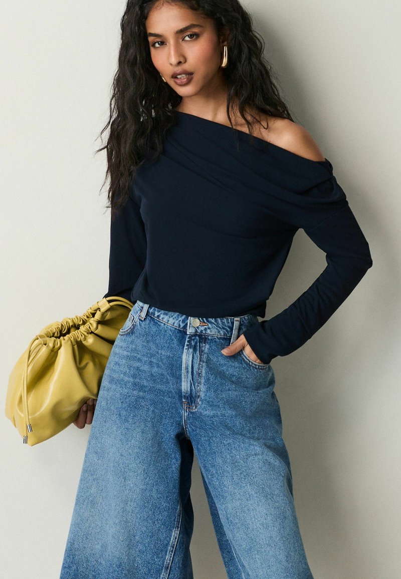 Námořnická off-shoulder blůza s dlouhými rukávy, spárovaná s vysokými, volně střiženými světle modrými džíny. Modelka drží žlutou sbíranou kabelku.