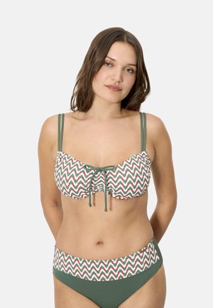 Mujer vistiendo un bikini con patrón de zigzag en verde y blanco, con doble tirante en los hombros y un detalle de lazo al frente, posando contra un fondo liso.