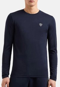 Maglietta a maniche lunghe di colore navy realizzata in tessuto liscio, con scollatura a girocollo e una piccola etichetta con logo sul lato sinistro del petto.