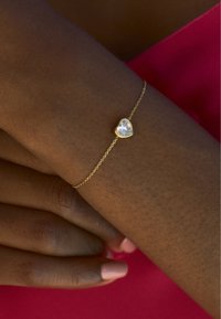 Ein goldfarbenes, herzförmiges Armband mit einem klaren Edelstein, der an einer zarten Kette befestigt ist. Die Hand hat einen hellen Hautton und trägt einen rosafarbenen Nagellack.