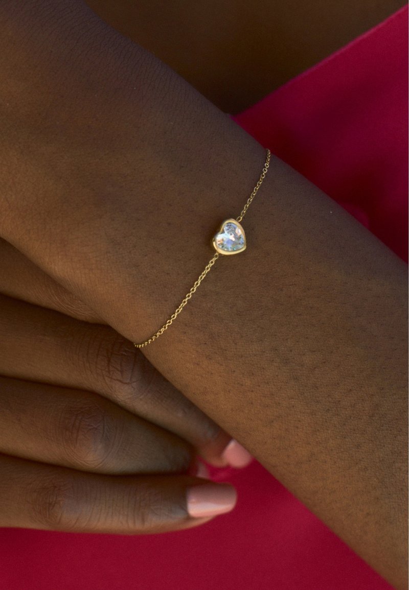 Ein goldfarbenes, herzförmiges Armband mit einem klaren Edelstein, der an einer zarten Kette befestigt ist. Die Hand hat einen hellen Hautton und trägt einen rosafarbenen Nagellack.