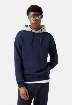 Mann trägt marineblauen Hoodie mit grauer Kapuze und großer verspiegelter Sonnenbrille, steht vor einem schlichten hellen Hintergrund.