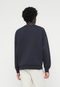 Personne aux cheveux courts et bouclés portant un sweat-shirt bleu marine foncé et un pantalon beige clair, vue de dos sur un fond clair uni.