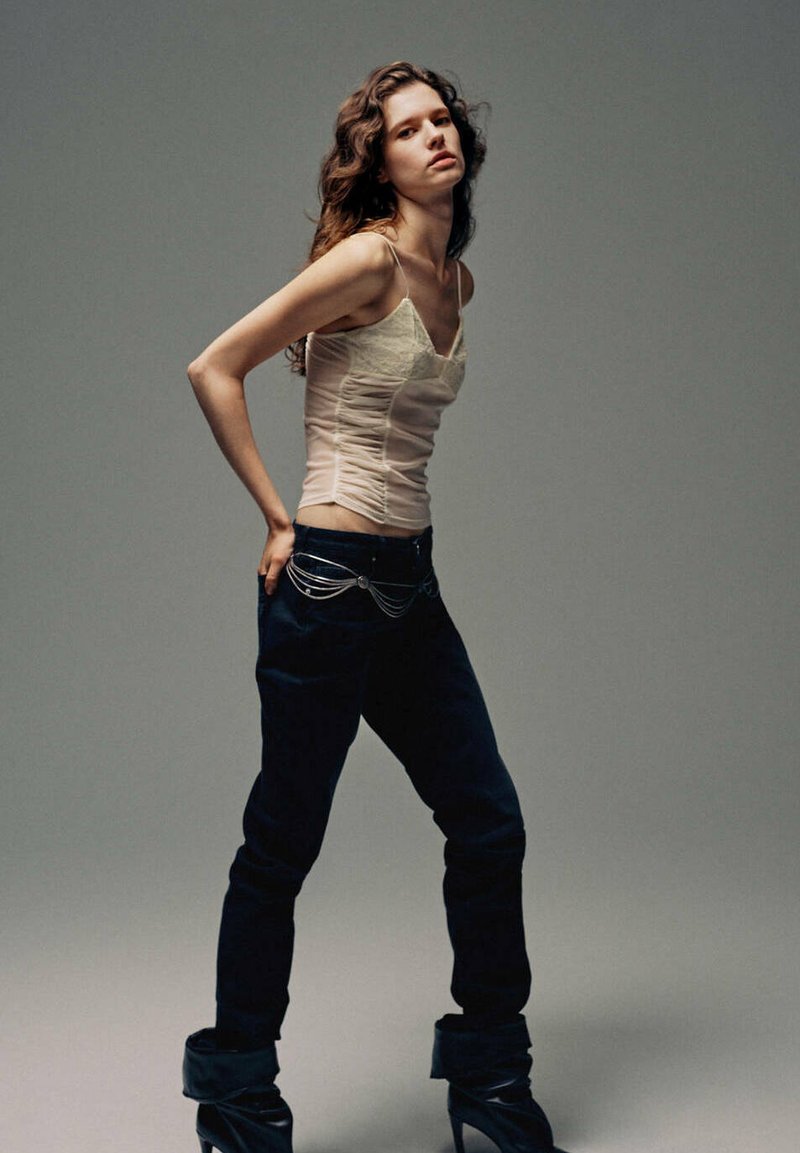 Jeune femme aux cheveux ondulés porte un caraco beige, un pantalon foncé avec une ceinture chaîne argentée et des bottines noires à talons, debout devant un fond uni.