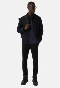 Marineblå uldblandet jakke med knapper, lagdelt over en lilla t-shirt; parret med marineblå bukser og sorte høje sneakers. Bærer en sort rygsæk.