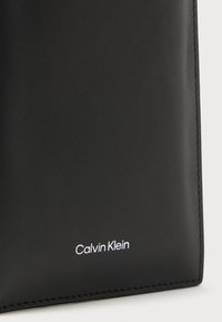 Juodas odinis piniginės, pasižyminčios lygiu paviršiumi, stačiakampio formos ir iškilniu balto spalvos "Calvin Klein" logotipu apačioje.