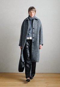 Garšs pelēks zīda mētelis ar melniem pogām, kas valkāts pār pelēku augsto kaklu un balto topu, kopā ar melnām mākslīgās ādas biksēm un apaviem.