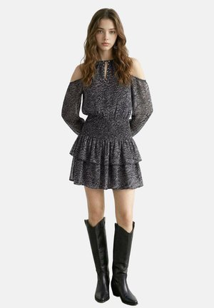 Young woman wearing a black patterned cold-shoulder dress with ruffled skirt and black knee-high boots, standing with hands behind back.