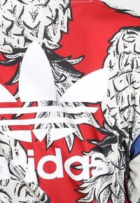 Logo Adidas z motywem trifolium w białym kolorze na abstrakcyjnie kwiecistym tle w czerwono-czarnych odcieniach.