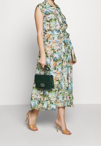 Blommönstrad, ärmlös klänning med bälte i midjan i lätt material; kombinerad med en grön handväska i krokodiltextur och nude klacksandaler.