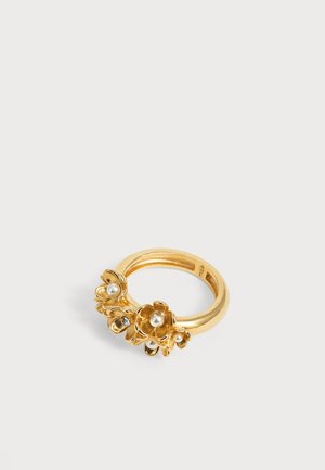 Bague en or avec trois petits motifs floraux, chacun avec une perle centrale et de petites pierres précieuses claires sur un fond blanc uni.