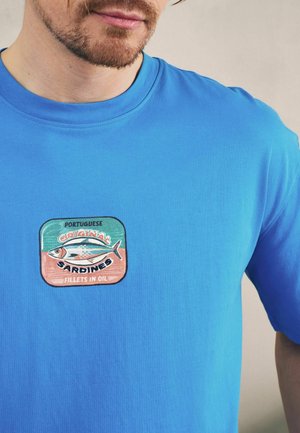 Mann trägt hellblaues T-Shirt mit einem bunten Aufnäher in Form einer Sardinendose mit der Aufschrift "Portuguese Original Sardines Fillets in Oil" auf der Brust.