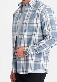 Camisa de manga larga a cuadros en azul y blanco, hecha de un tejido suave, con un bolsillo en el pecho y puños abotonados, superpuesta sobre una camiseta gris.