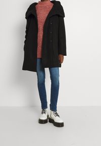 Persoon in een zwarte middellange jas, een rode gebreide trui, blauwe skinny jeans en witte platform veterschoenen staat tegen een witte achtergrond.