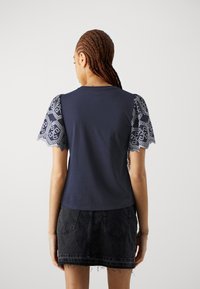 Kvinna med långt flätat hår som bär en marinblå topp med mönstrade korta ärmar och en svart jeanskjol, vänd bort mot en vit bakgrund.