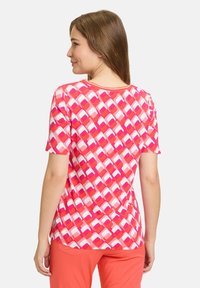 Kortærmet lyserød t-shirt med et geometrisk mønster af firkanter i varierende nuancer, glat tekstur og rund hals. Båret med koralfarvede bukser.