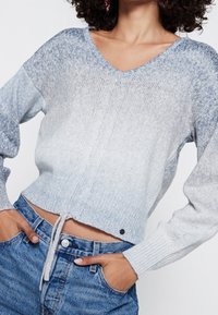 Pull court bleu clair en maille, avec un col en V, des manches longues et un détail de cordon à la base. Motif dégradé texturé.