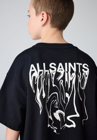 Poiss, kes on küljega, kannab musta üle suurusega T-särki, millel on taga "ALLSAINTS" tekst ja valge moonutatud graafiline kujundus.