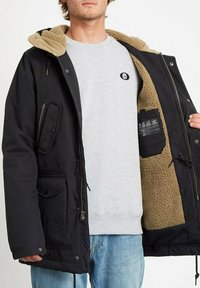Schwarze Jacke mit einem tanfarbenen Fleece-Innenfutter, mit einem Reißverschluss und Knopfverschluss an der Vorderseite, mehreren Taschen und einem Kordelzug am Saum.