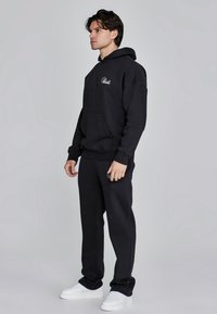 Zwarte oversized hoodie met voorzak en klein wit logo, gecombineerd met bijpassende zwarte joggingbroek en witte sneakers.