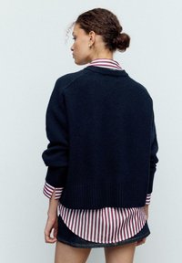 Temno modra pletena majica z ohlapnim krojem in rebrastimi manšetami, nošena preko bele srajce z bordo rdečimi črtami, s ukrivljenim robom.