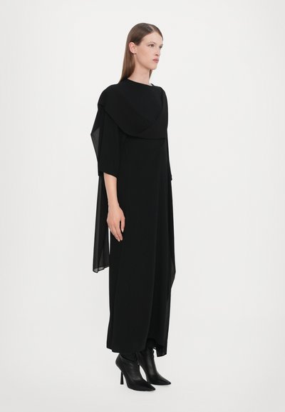 Black long dress with a draped collar and sheer accents, featuring three-quarter sleeves and a relaxed silhouette. Worn with black heeled boots.